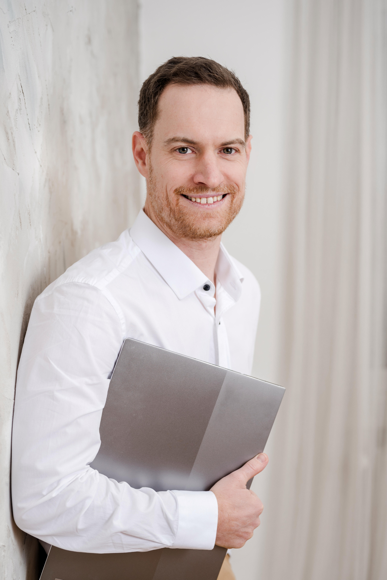 Personal Branding Fotografie Wien: Markus Schick lächelt im weißen Hemd mit einem Laptop unter dem Arm vor einem hellen, neutralen Hintergrund.